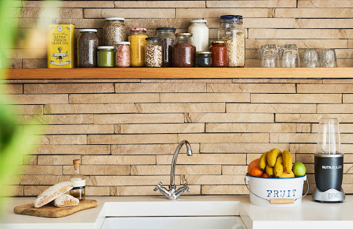 white countertop displaying blender, cutting board with bread, and a tin of fruit with a sink in front of a natural wooden backsplash and a wooden shelf with spices above